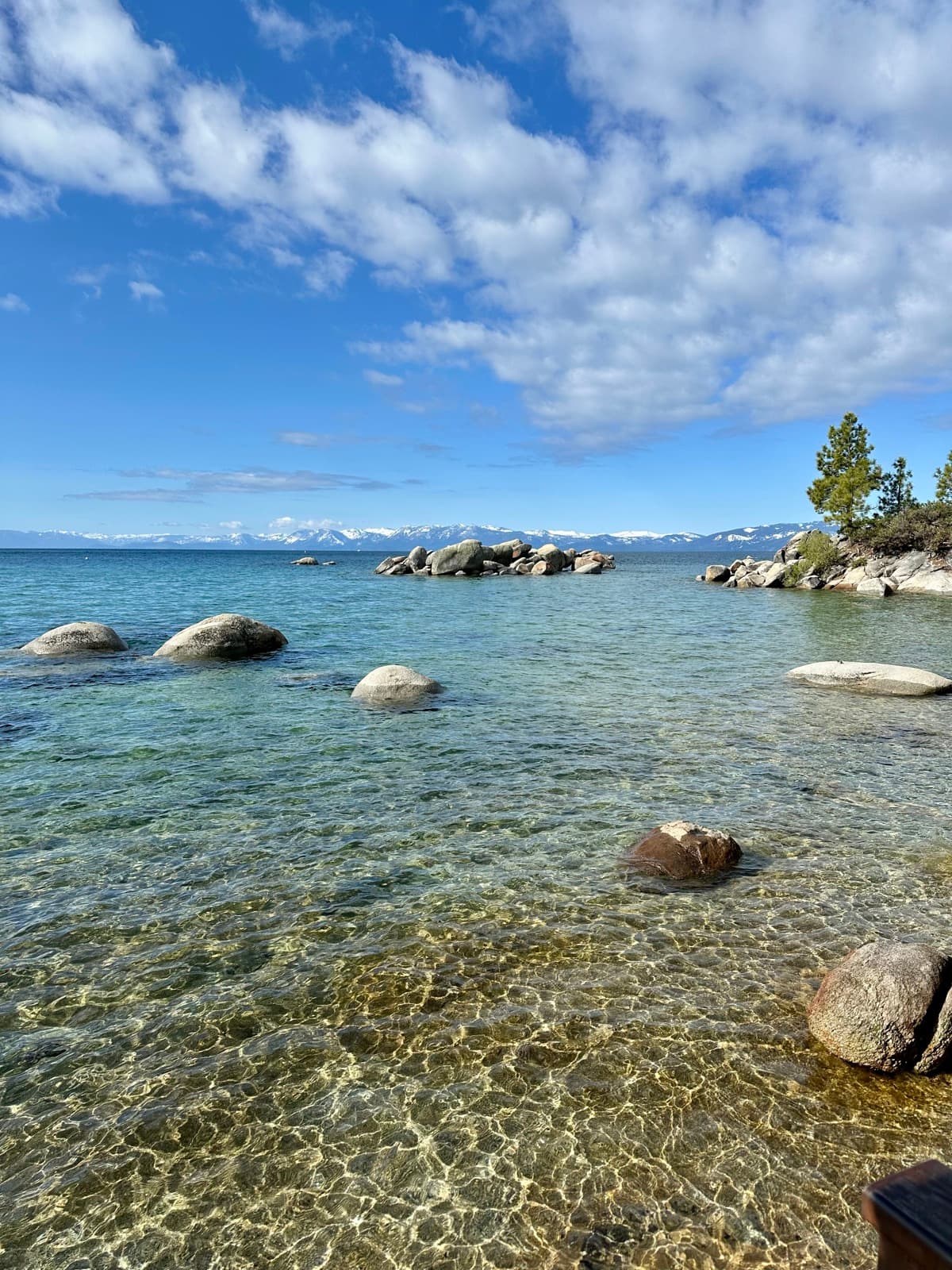 A clear alpine lake shore with boulders and snow-capped mountains beyond